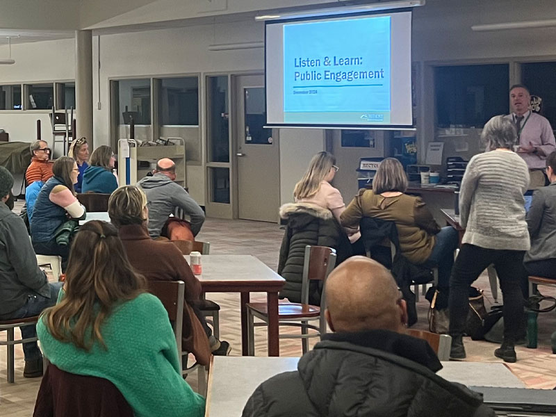 A Listen and Learn public Engagement session at CVU. Members of the public sit and listen to a presentation by Agency staff. There is a screen displaying a slide that says "Listen & Learn: Public Engagement."