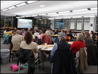 30 adults sit at tables in a school library watching a slide presentation on a large screen.